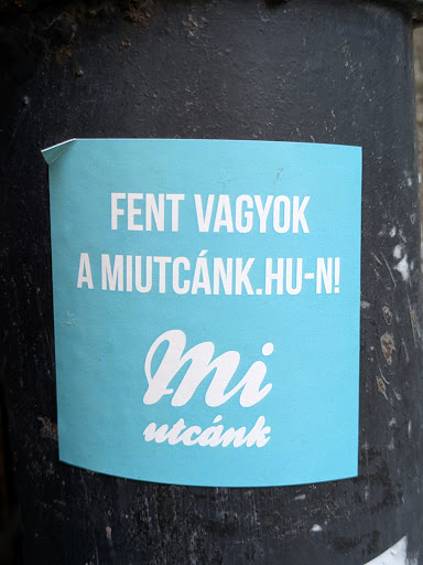 Street sticker A light teal square sticker with white text on a dark gray surface. The text says "Fent vagyok a miutcánk.hu-n!" which translates to "I'm on miutcánk.hu!" in English. There is also a stylized "Mi utcánk" logo.