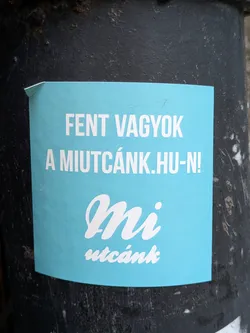 Street sticker A light teal square sticker with white text on a dark gray surface. The text says "Fent vagyok a miutcánk.hu-n!" which translates to "I'm on miutcánk.hu!" in English. There is also a stylized "Mi utcánk" logo.