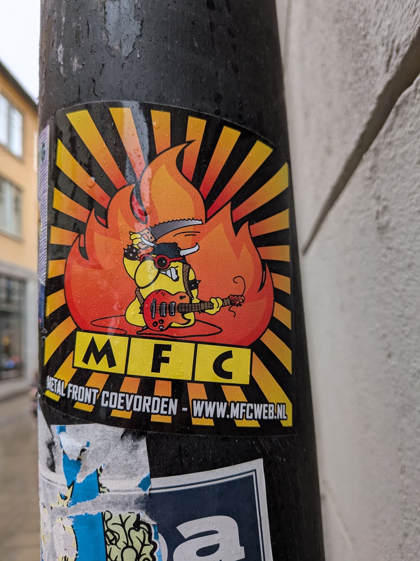 A vibrant, rectangular sticker featuring a cartoonish yellow creature with a large mouth, red sunglasses, a black horned helmet, and a spiked wristband, passionately playing a red electric guitar. The creature is engulfed in large, stylized orange and red flames. The background consists of a radial sunburst pattern in alternating black and yellow/orange stripes. Below the main illustration, the letters 'M F C' are prominently displayed in yellow blocks with black outlines. Further below, in white text, are 'METAL FRONT COEVORDEN' and 'WWW.MFCWEB.NL'. The sticker has a glossy finish and shows visible water droplets, indicating it's outdoors.