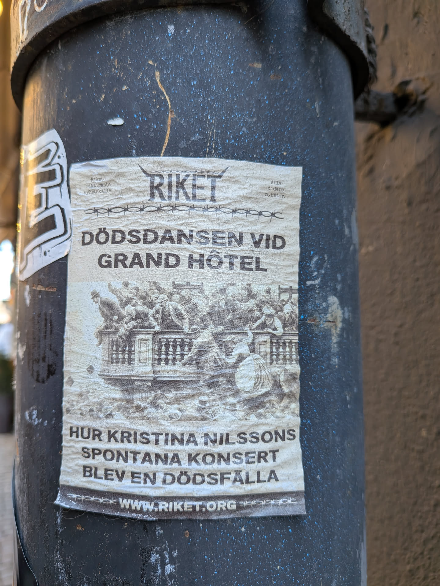 The sticker is a rectangular, off-white paper, heavily creased and worn, affixed to a dark, textured pole. At the top, the word 'RIKET' is prominently displayed in a bold, dark font, flanked by two horn-like symbols. Below this, a decorative line resembling barbed wire is visible. The main title, 'DÖDSDANSEN VID GRAND HÔTEL', is written in large, capitalized letters. Below the title, a detailed, monochrome illustration depicts a chaotic historical scene on a balcony or balustrade, with numerous figures in period attire appearing to fall or struggle amidst a crowd. Further down, the text 'HUR KRISTINA NILSSONS SPONTANA KONSERT BLEV EN DÖDSFÄLLA' is printed. Small text 'Rikets riktigaste nyhetskälla.' is in the top left, and 'Alla tiders nyheter.' in the top right. At the very bottom, the website 'WWW.RIKET.ORG' is clearly visible. The overall aesthetic is vintage and dramatic, reflecting a historical news or event announcement.
