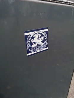 Street sticker The sticker features a striking design in dark blue and white. At its center is a white, rearing Pegasus with prominent wings, enclosed within a circular white wreath-like border. Below the Pegasus, a white banner unfurls, displaying the text "B*BLOCK" on the left and "STUTTGART" on the right, separated by a small star. The entire central emblem is set against a dark blue background, which is framed by horizontal white stripes at the top and bottom, giving it a stylized flag or banner appearance. The sticker has slightly rounded corners and is applied to a dark, possibly metallic, surface.