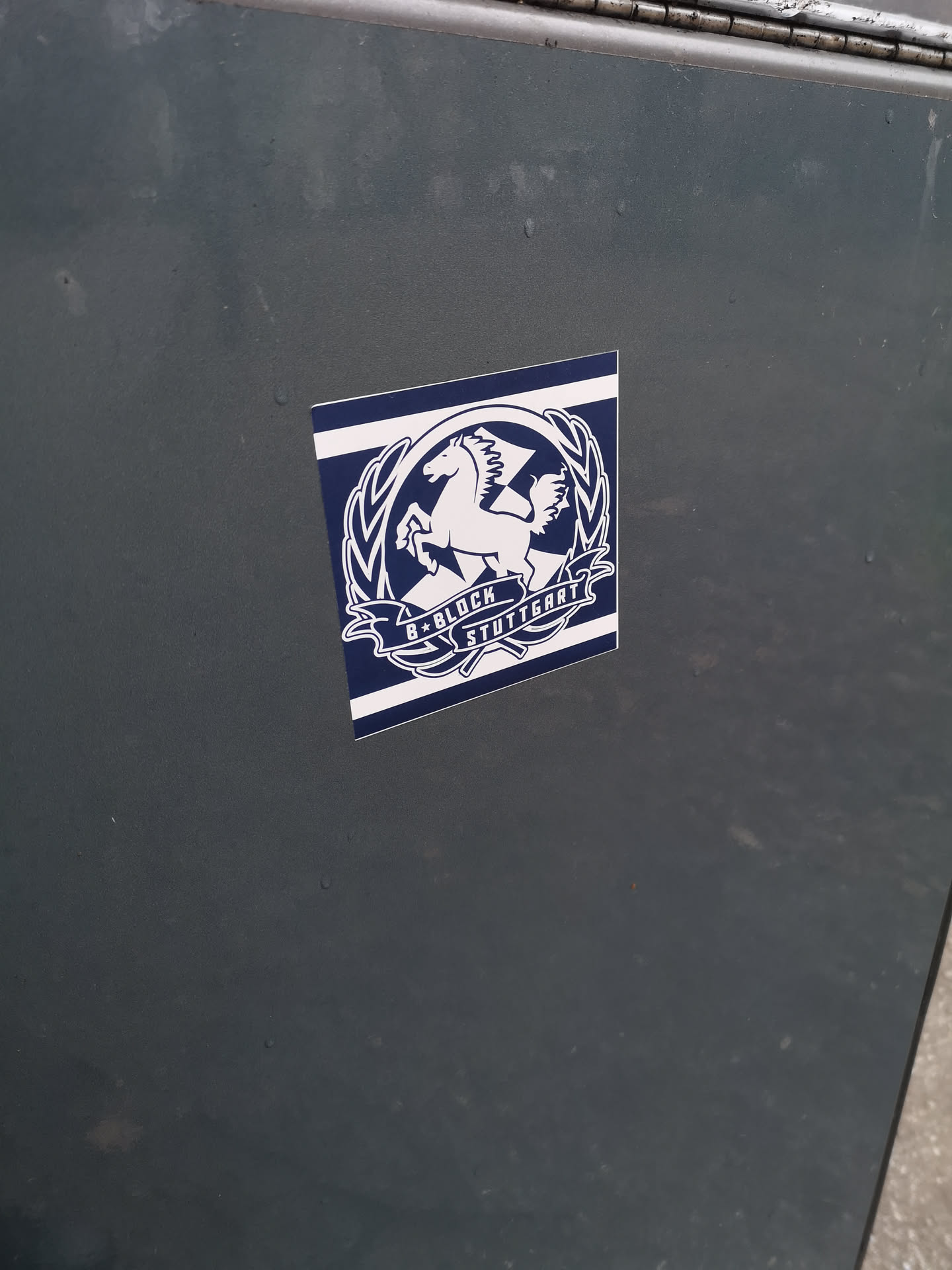 The sticker features a striking design in dark blue and white. At its center is a white, rearing Pegasus with prominent wings, enclosed within a circular white wreath-like border. Below the Pegasus, a white banner unfurls, displaying the text "B*BLOCK" on the left and "STUTTGART" on the right, separated by a small star. The entire central emblem is set against a dark blue background, which is framed by horizontal white stripes at the top and bottom, giving it a stylized flag or banner appearance. The sticker has slightly rounded corners and is applied to a dark, possibly metallic, surface.