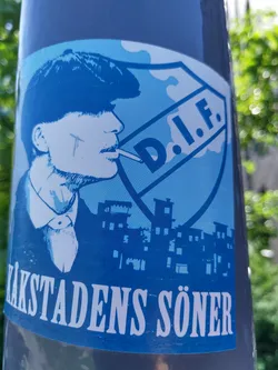 Street sticker A blue and white sticker featuring a stylized illustration of a man resembling Tommy Shelby from the Peaky Blinders TV series.  He is smoking a cigarette and is set against a backdrop of buildings, with a prominent shield-shaped logo reading "D.I.F." The bottom of the sticker displays the text "ÄKSTADENS SÖNER" in white lettering.