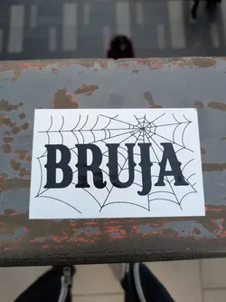 Street sticker A rectangular sticker featuring the word "Bruja" in a bold, gothic-style font. The word is laid over a stylized spiderweb design. The sticker is placed on a weathered metallic surface.