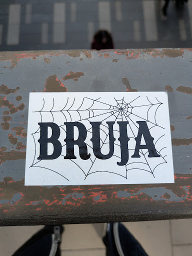 A rectangular sticker featuring the word "Bruja" in a bold, gothic-style font. The word is laid over a stylized spiderweb design. The sticker is placed on a weathered metallic surface.