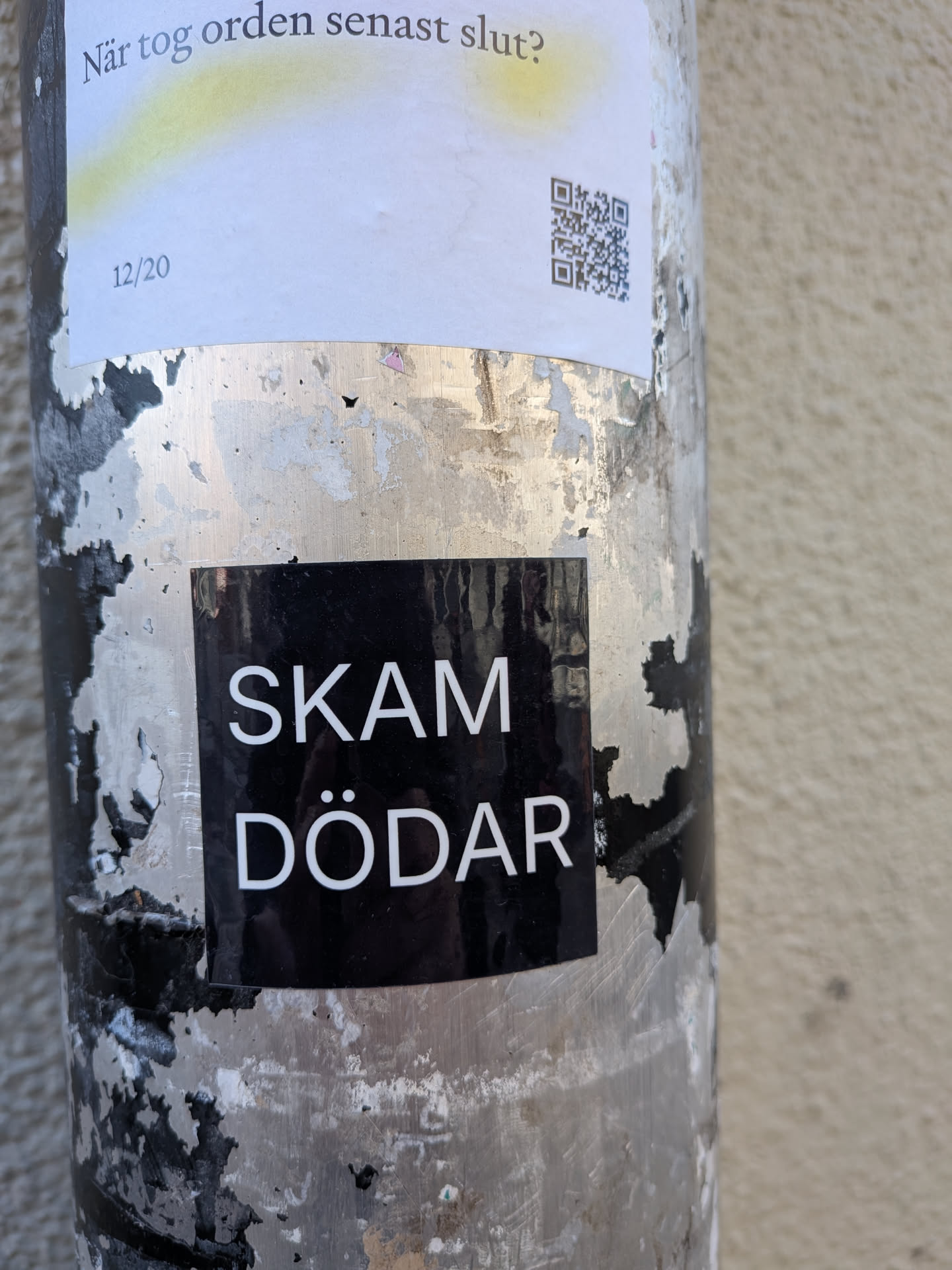 The sticker is a rectangular, black background with two lines of bold, white, sans-serif text. The top line reads 'SKAM' and the bottom line reads 'DÖDAR'. The sticker has a slight sheen, indicating a vinyl or similar material, and is applied to a heavily worn, metallic pole with peeling paint. The edges of the sticker appear crisp and well-defined.