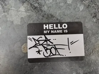 Street sticker A black and white sticker resembling a name tag with the text "HELLO MY NAME IS" printed on it.  It has been customized with black graffiti art drawn over the name area. The sticker is affixed to a weathered metallic surface.