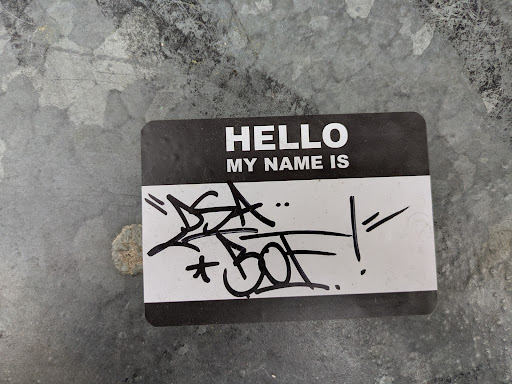 A black and white sticker resembling a name tag with the text "HELLO MY NAME IS" printed on it.  It has been customized with black graffiti art drawn over the name area. The sticker is affixed to a weathered metallic surface.