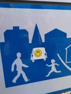 Street sticker The sticker features a universally recognized yellow smiley face. It consists of a bright yellow circular background with two simple black oval eyes and a wide, curved black line forming a smile. The smiley face is printed on a small, roughly rectangular white sticker, which is slightly creased and appears to be manually applied to a larger blue and white public sign. The edges of the white sticker are visible, indicating it's a separate adhesive element.