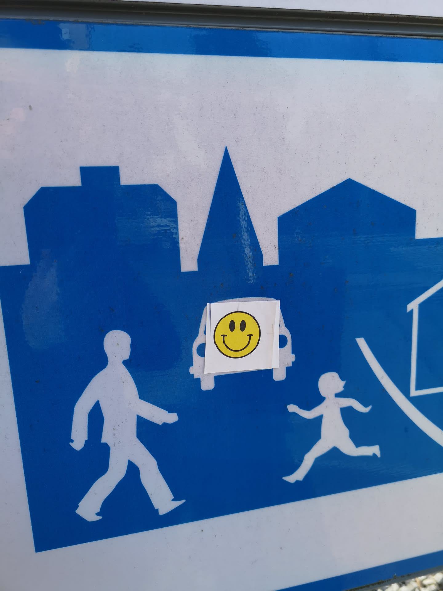 The sticker features a universally recognized yellow smiley face. It consists of a bright yellow circular background with two simple black oval eyes and a wide, curved black line forming a smile. The smiley face is printed on a small, roughly rectangular white sticker, which is slightly creased and appears to be manually applied to a larger blue and white public sign. The edges of the white sticker are visible, indicating it's a separate adhesive element.