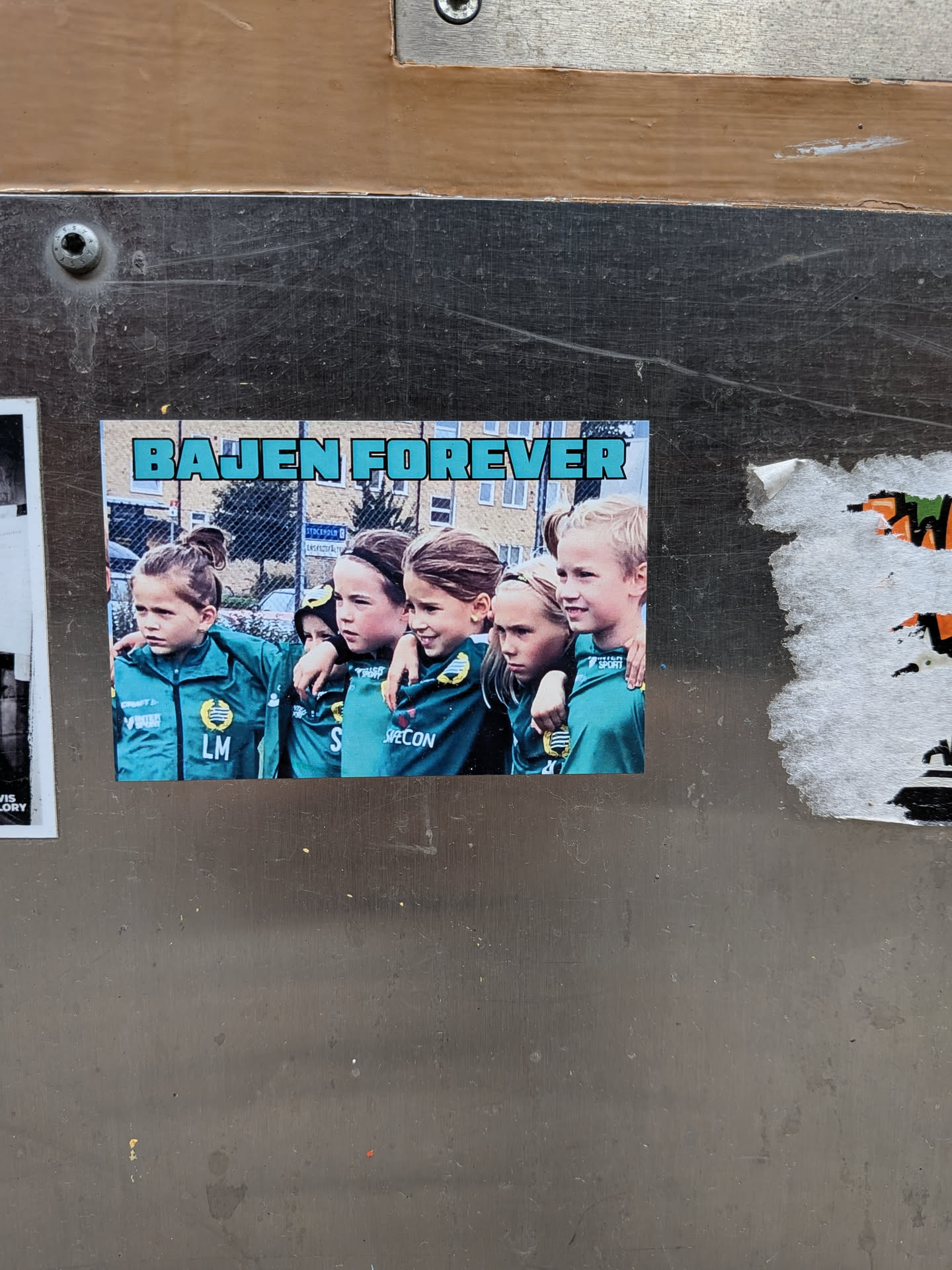 A rectangular sticker featuring a photograph of five young girls, likely a youth soccer team, standing close together with their arms around each other. They are all wearing matching teal-green long-sleeved soccer jerseys with yellow and black club crests on the left chest. Various sponsor logos like 'CRAFT', 'VINTER', 'TRUST SPORT', 'S', and 'SPECON' are visible on their jerseys, along with initials like 'LM'. The girls have diverse hair colors and styles, and they appear to be smiling or looking forward with determination. Above the photo, in bold, blocky, light blue letters with a dark outline, is the text 'BAJEN FOREVER'. The background of the photo shows blurred buildings and a chain-link fence, with a street sign partially visible that reads 'STOCKHOLM'. The sticker itself is adhered to a scratched, metallic surface.