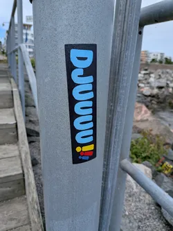 Street sticker A vertical black rectangular sticker on a metal post. It displays the word 'DJUUUU' in rounded light blue letters, ending with three exclamation marks in yellow, red, and blue.