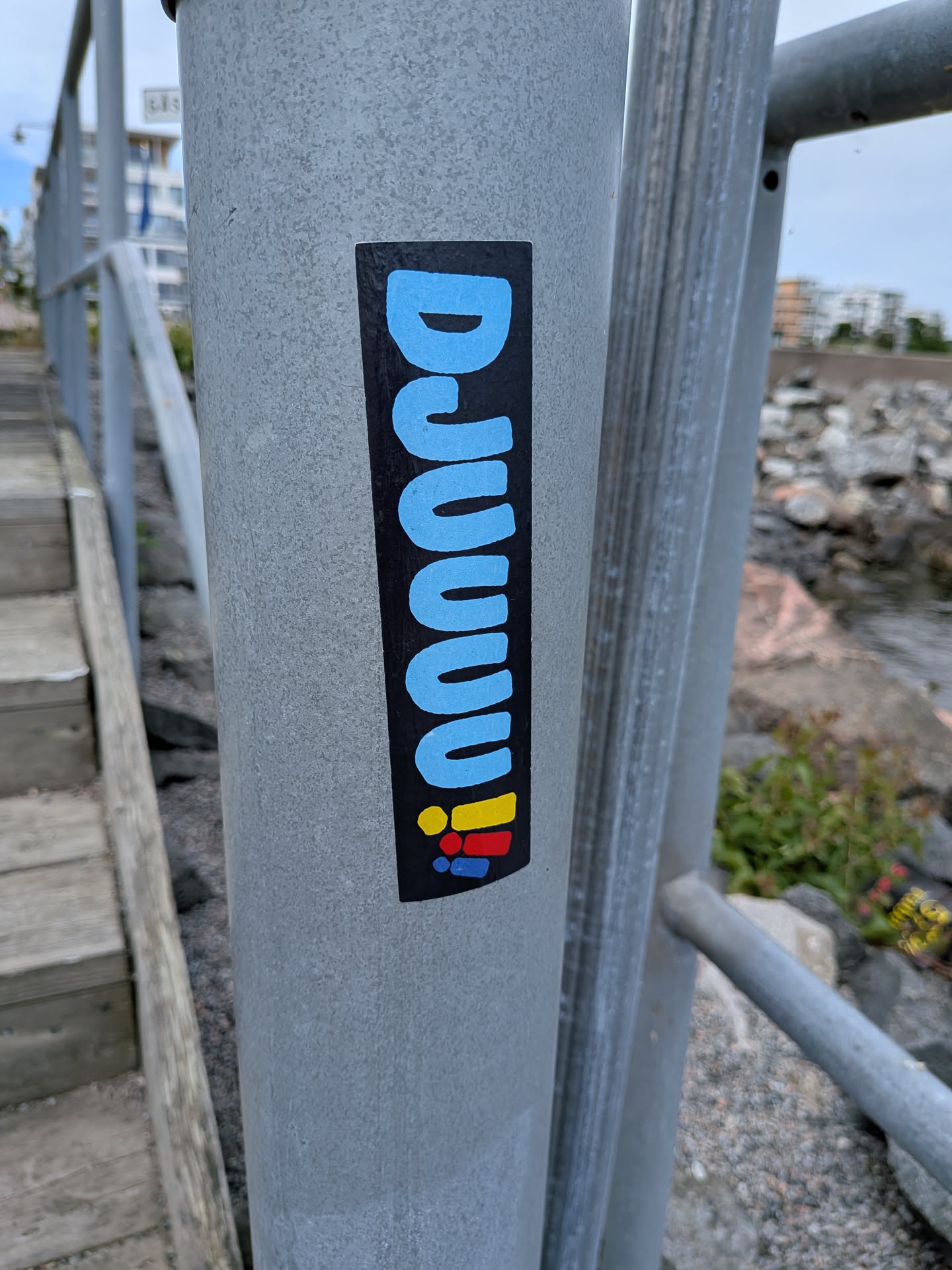 A vertical black rectangular sticker on a metal post. It displays the word 'DJUUUU' in rounded light blue letters, ending with three exclamation marks in yellow, red, and blue.