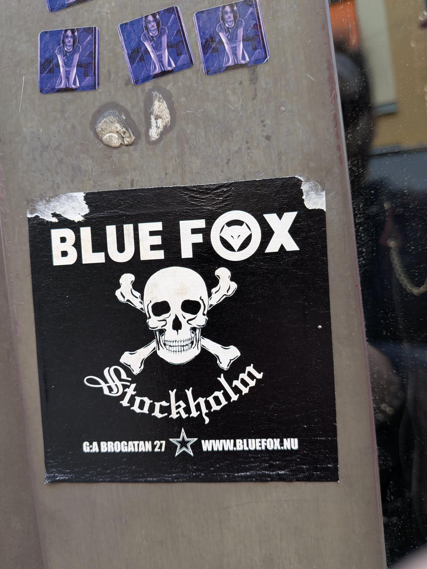 A square black sticker featuring white text and graphics. It displays 'BLUE FOX' with a fox head icon, a central skull and crossbones, 'Stockholm' in a gothic font, and store address and website details at the bottom.