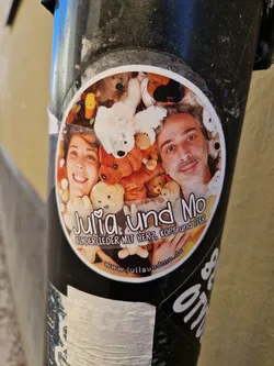 A round sticker featuring Julia and Mo, surrounded by stuffed animals.  The text on the sticker indicates they make children's songs. A website address is also included.