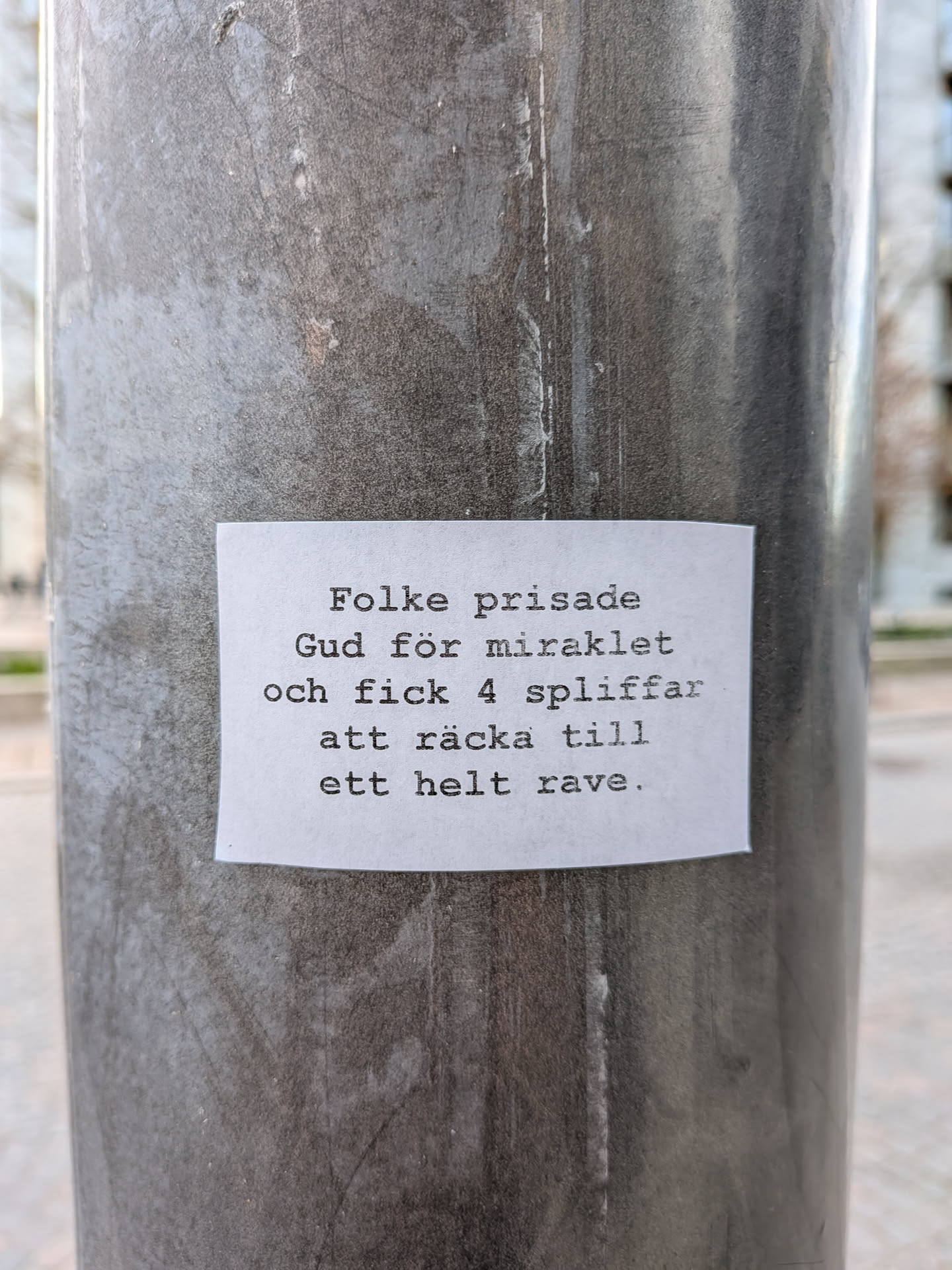 A rectangular white paper sticker with black text in a typewriter-style font, adhered to a textured, dark grey metal pole. The sticker appears slightly worn with minor imperfections and edges that suggest it's a physical object stuck to the surface. The text is centrally aligned on the sticker.