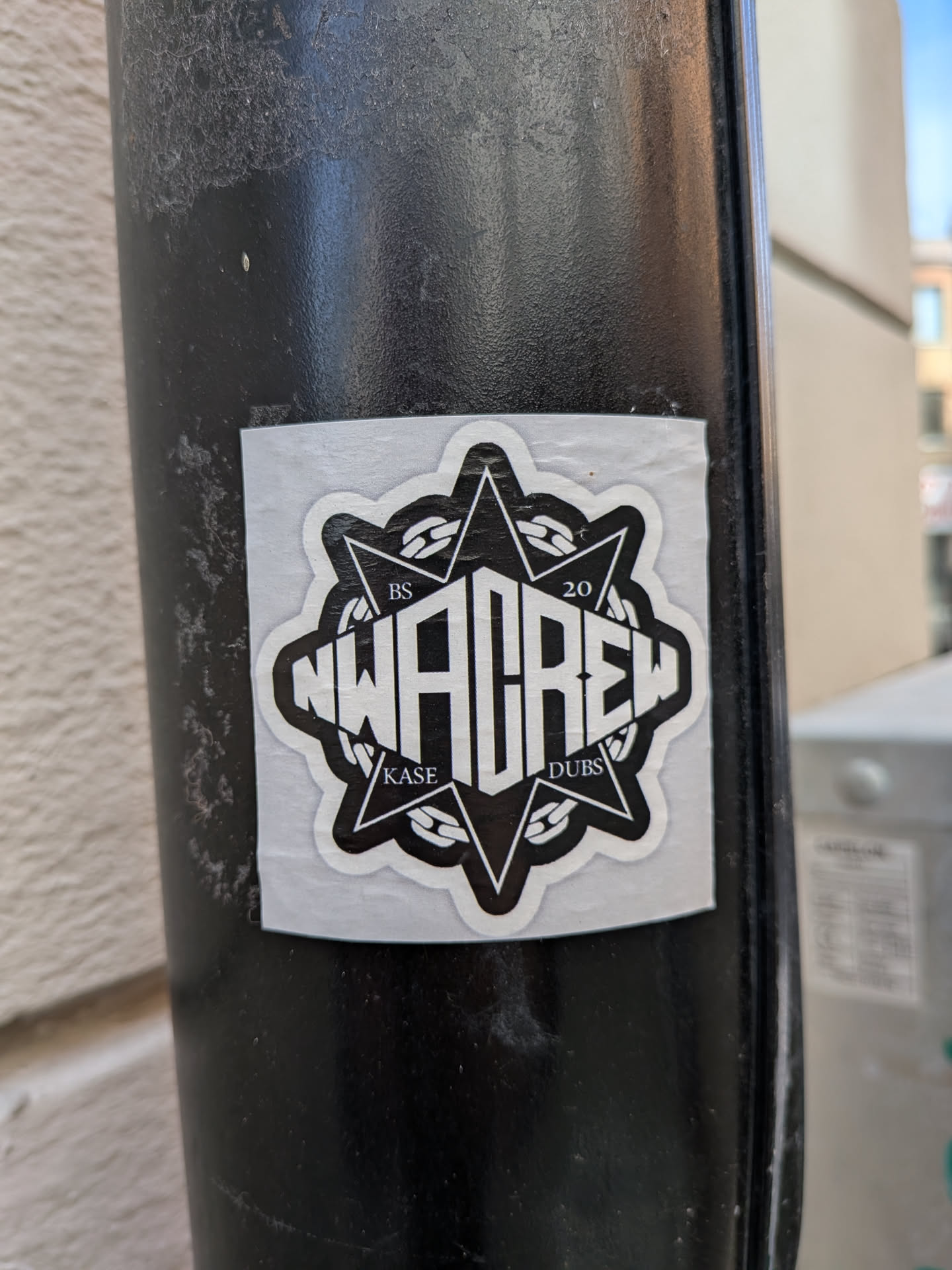 A square sticker with a black circular emblem on a white background. The emblem features stylized white text reading 'NWA CREW' across the center, set against an eight-pointed star and a circular chain motif. Small text reads 'BS', '20', 'KASE', and 'DUBS' in the star's corners.