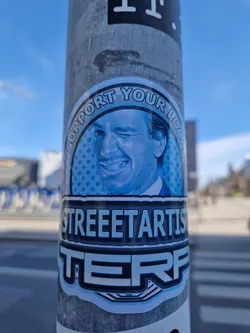 Street sticker A blue and white sticker featuring a winking man's face in a circle with the text 'Support Your Local' above and 'STREETARTISTERF' below. The sticker has a pop art style.