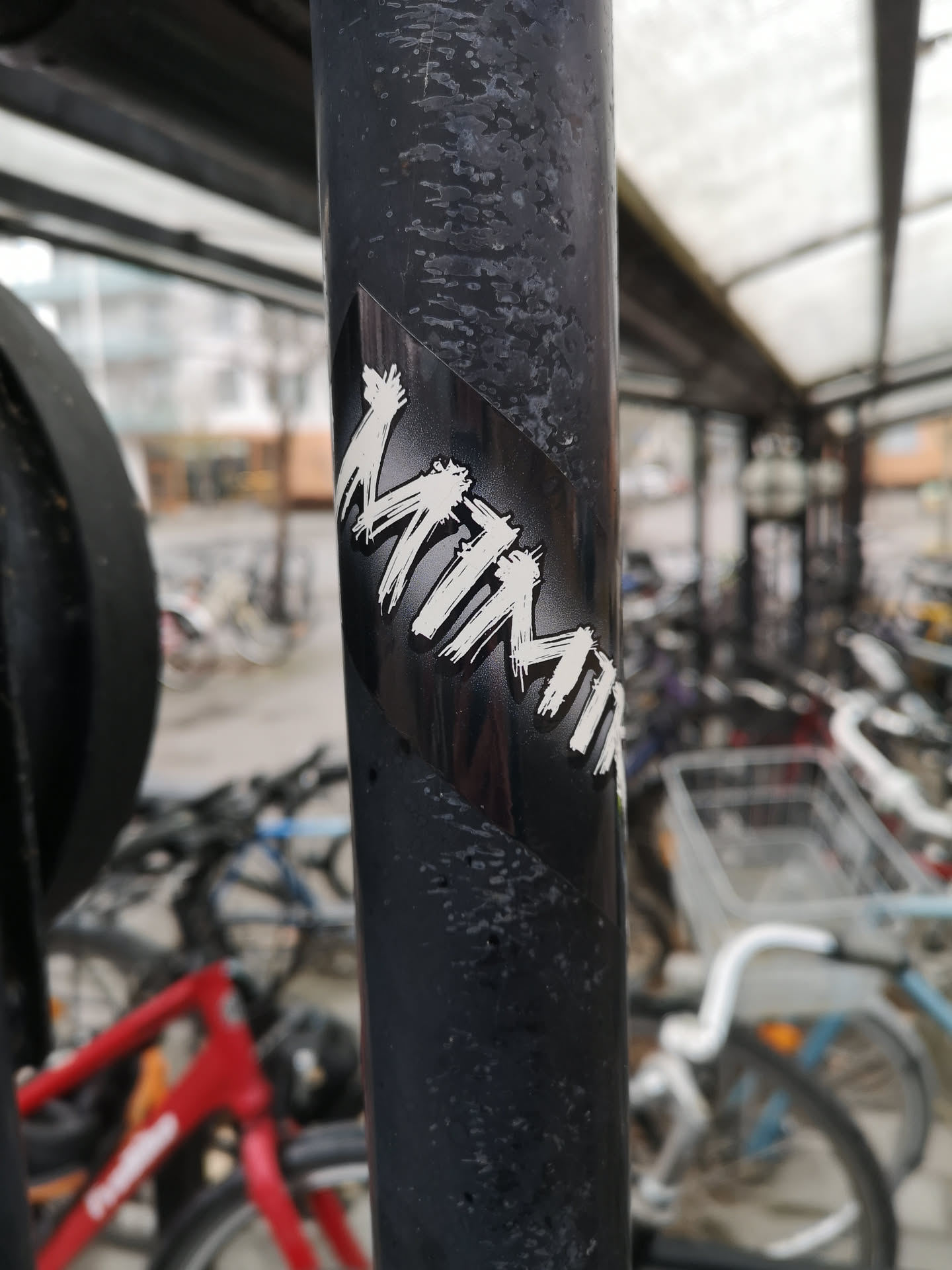 The sticker is rectangular, featuring the word "MIMMI" in a prominent, white, brushstroke-style font against a solid black background. The letters have a dynamic, hand-painted appearance with visible texture, suggesting a raw, energetic aesthetic. The sticker is applied diagonally on a dark, textured metal pole, contrasting sharply with its surroundings.