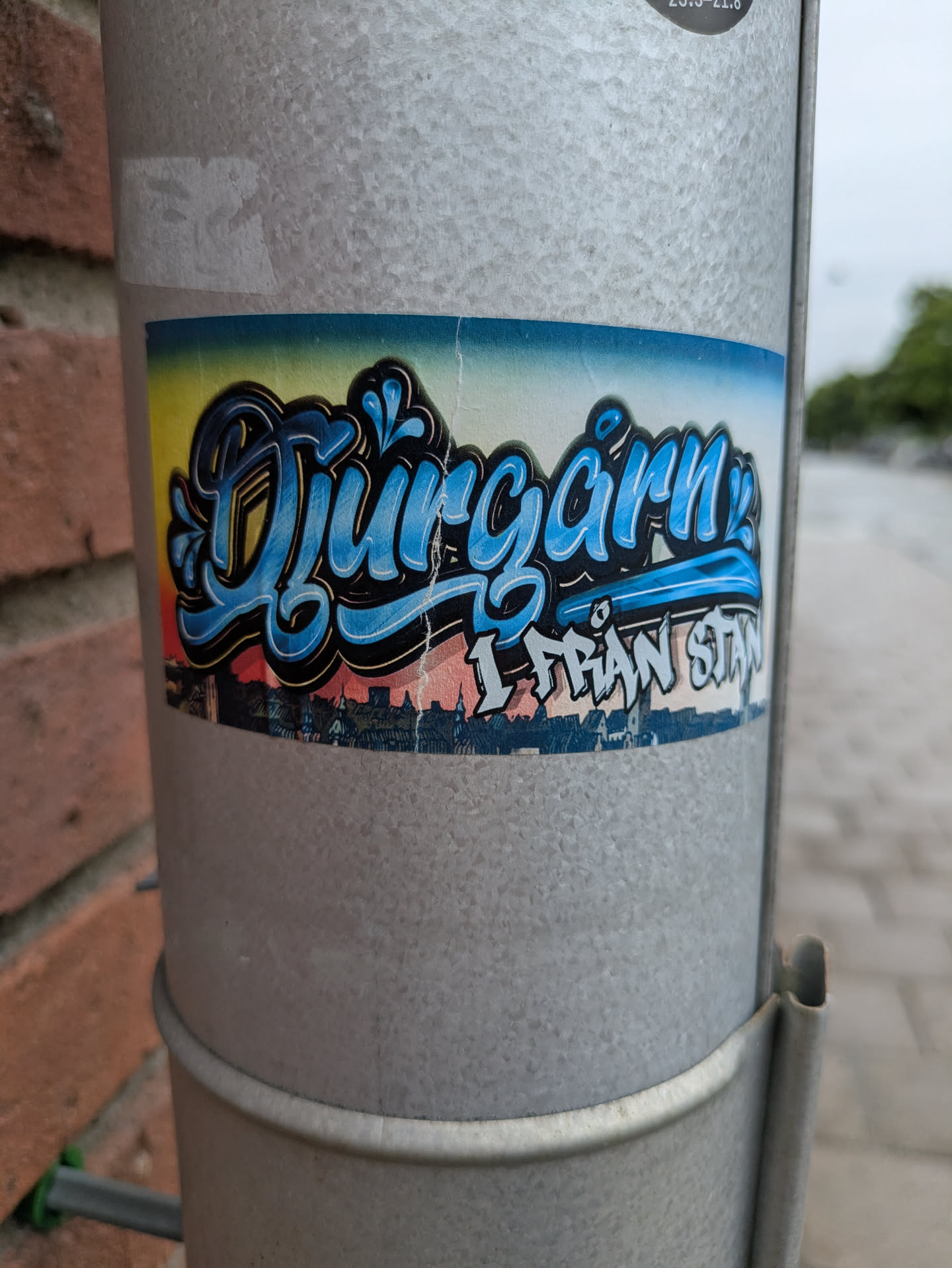 The sticker is rectangular, featuring the word "Djurgårn" prominently in large, light blue bubble letters with a dark blue and black outline, rendered in a graffiti style. Below it, in smaller, blocky white letters with a black outline, is the phrase "I FRÅN STAN". The background behind the text is a gradient sky, transitioning from yellow on the left to light blue in the middle, and then to a pinkish-orange hue on the right, resembling a sunset or sunrise. Beneath this sky, a dark silhouette of a city skyline with various buildings is visible. The sticker is applied to a textured, galvanized metal pole, showing some wear and slight creasing.