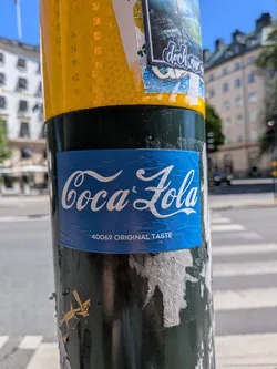 Street sticker The sticker is a rectangular blue label with white text, mimicking the iconic Coca-Cola logo. The main text, in a flowing script font, reads 'Coca & Zola'. Below this, in a smaller, sans-serif font, it says '40069 ORIGINAL TASTE'. The sticker is adhered to a dark pole and shows some wrinkles and minor wear, indicating it's a physical sticker in an urban environment. The background of the sticker is a solid, medium-dark blue.