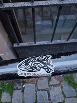 Street sticker A sticker featuring a minimalist line art drawing of a heart. The sticker is attached to a black metal railing outdoors.  The text 'Gapa Studios.se' is visible at the bottom of the sticker.
