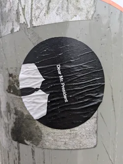 Street sticker This round sticker features a stark contrast of black and white. The text 'Dear Mr. President' is printed along the right side of the sticker. To the left of the text is an abstract image of a broken white tie set against the black background. The sticker is adhered to a gray textured surface.
