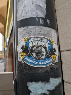 Street sticker The sticker is a shield-shaped emblem with a prominent white outline, featuring a dynamic design in blue, black, white, and yellow. The top banner, set against a blue background, boldly declares "FRIHET FÖR ULTRAS" (Freedom for Ultras) in white block letters. The central part of the shield contains a stylized black and white graphic, possibly depicting a figure or symbol related to fan culture, with a large 'S' or '5' in the background. Flanking the central shield on both sides are white megaphones with black interiors, outlined in black, and adorned with yellow laurel-like leaves. A lower blue banner, mirroring the top, reads "SYMPATI FÖR DE AVSTÄNGDA" (Sympathy for the Banned) in white block letters. The overall aesthetic is reminiscent of a sports club crest or a fan group's official logo, conveying a strong message of support and defiance.