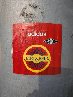 Street sticker The sticker is predominantly red with vertical stripes. At the top, the white Adidas logo is clearly visible. To the right of the Adidas logo, there is a black and white diamond shape with the text '19 RBK 17' inside. The center of the sticker features a large yellow circle with a dark blue outline. Inside this circle are stylized blue floral or leaf-like shapes. Below these shapes, the word 'JARLSBERG' is written in bold, dark blue capital letters, with a small registered trademark symbol to its right.