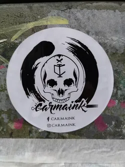 Street sticker Round white sticker with a black and white illustration of a skull inside an enso circle. The skull has a symbol on its forehead.  Below the artwork, the text 'Carmaink' is written in a stylized script font. Social media handles for Facebook and Instagram are also present.