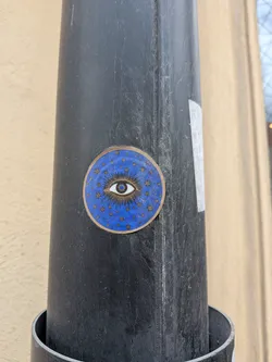 Street sticker The sticker is circular with a prominent gold-colored border. Its background is a deep, rich blue. At the center, a stylized eye is depicted in white with a blue iris and a dark pupil. Numerous thin, gold lines radiate outwards from the eye, creating a sunburst effect. Scattered across the blue background, beyond the immediate rays, are many small, gold four-pointed stars. The sticker has a slightly worn appearance, consistent with being affixed outdoors.