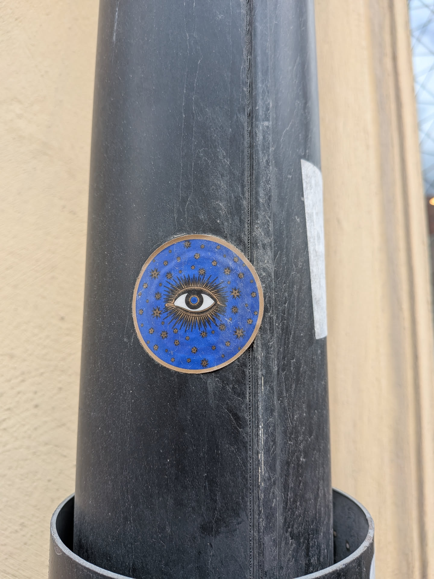 The sticker is circular with a prominent gold-colored border. Its background is a deep, rich blue. At the center, a stylized eye is depicted in white with a blue iris and a dark pupil. Numerous thin, gold lines radiate outwards from the eye, creating a sunburst effect. Scattered across the blue background, beyond the immediate rays, are many small, gold four-pointed stars. The sticker has a slightly worn appearance, consistent with being affixed outdoors.