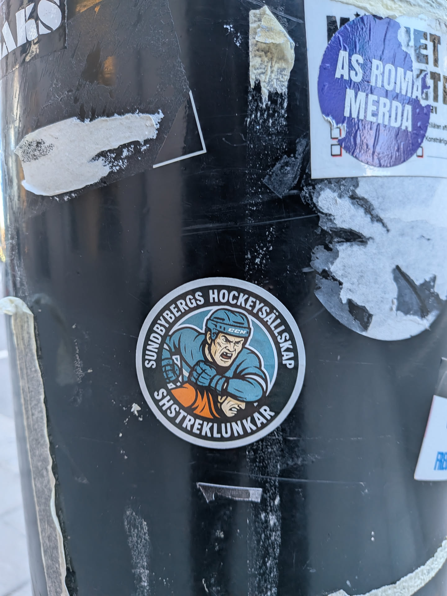 A circular sticker featuring a stylized, aggressive hockey player. The player is depicted from the waist up, wearing a light blue helmet with a 'CCM' logo, a light blue jersey, and orange gloves. His face shows a grimace with bared teeth and intense eyes, conveying a sense of determination or aggression. The background behind the player is a lighter blue circle. The sticker has a white and black border, with text arched along the top and bottom edges. The overall color scheme includes light blue, orange, black, white, and skin tones.