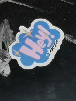 Street sticker The sticker is shaped like a stylized, puffy cloud or speech bubble, outlined in white. The main body of the cloud is a vibrant sky blue. Inside, the word "Hey!" is written in a bold, rounded, light pink font. Three small, four-pointed golden-yellow stars are scattered around the text within the blue cloud, adding a sparkling effect. In the bottom right corner, outside the main blue cloud but within the white outline, the word "vistaprint" is written vertically in black, accompanied by a small blue and white triangular logo. The sticker appears to be die-cut, following its irregular cloud shape, and is placed on a dark, possibly black or dark gray, textured surface.