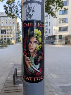 Street sticker The sticker prominently displays a stylized portrait of a woman with piercing blue eyes, dark wavy hair intertwined with vibrant green leaves, and delicate facial features. She is depicted holding a gleaming, sharp knife. The background transitions from a fiery red glow behind her head to black. The text 'EMILLIOS' is arched above her head in bold, red lettering with a yellow outline, and 'TATTOO' is displayed below her chin in similar, but slightly smaller, yellow lettering with a red outline. The overall style evokes a traditional tattoo aesthetic.