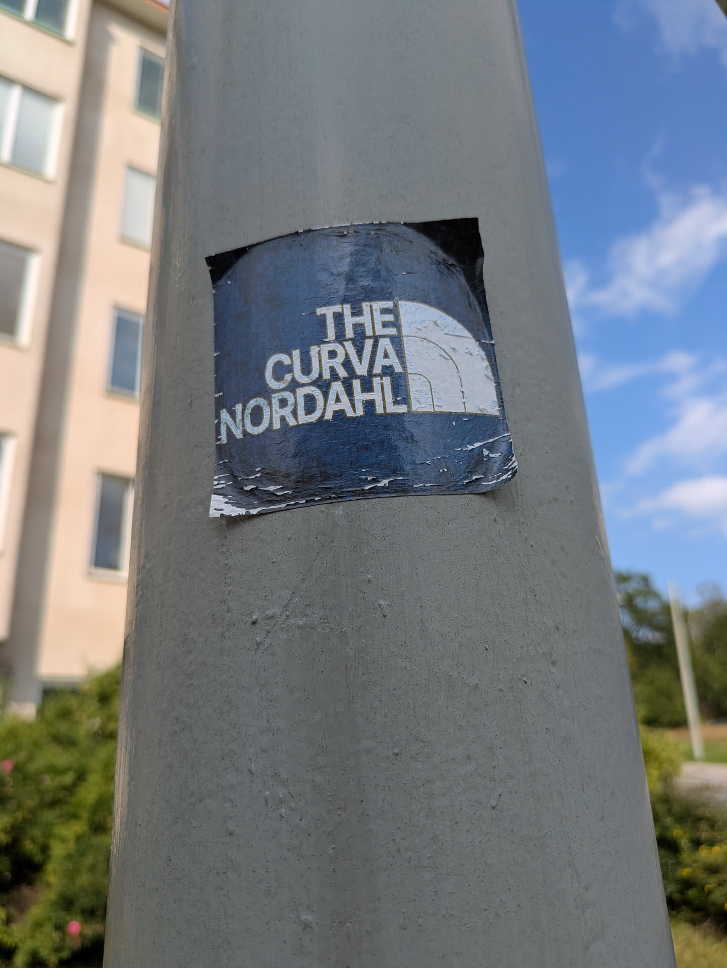 The sticker is a roughly square-shaped design with a dark blue or navy background. On the right side, there is a prominent white graphic, which is a clear parody of The North Face's half-dome logo. To the left of this graphic, white, sans-serif text is arranged in two lines. The top line reads 'THE', and the bottom line reads 'CURVA NORDAHL'. The sticker shows significant signs of wear and tear, with peeling edges, visible creases, and scuffs across its surface, indicating exposure to the elements. It is adhered to a light grey, cylindrical pole.