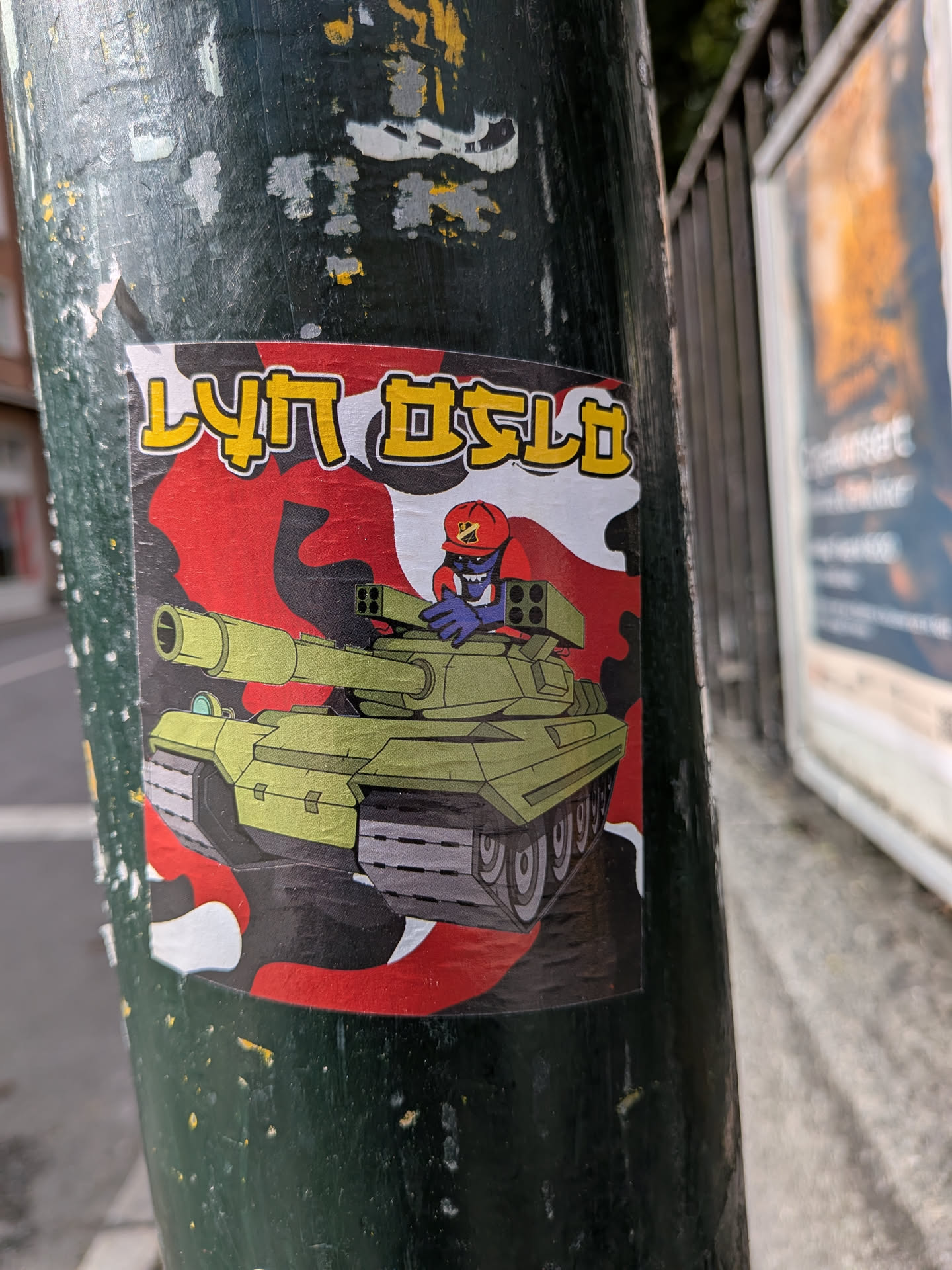 The sticker features a cartoonish illustration of a light green tank with a red and black camouflage pattern in the background. A blue creature with a red cap and a determined expression is depicted in the driver's seat, operating the tank's mounted weapon. The text 'LYN OSLO' is prominently displayed in yellow capital letters with a black outline at the top of the sticker.