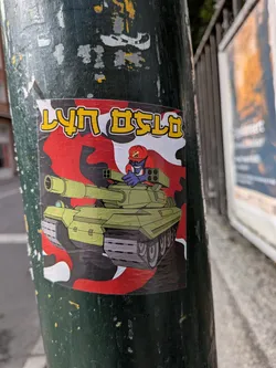 Street sticker The sticker features a cartoonish illustration of a light green tank with a red and black camouflage pattern in the background. A blue creature with a red cap and a determined expression is depicted in the driver's seat, operating the tank's mounted weapon. The text 'LYN OSLO' is prominently displayed in yellow capital letters with a black outline at the top of the sticker.