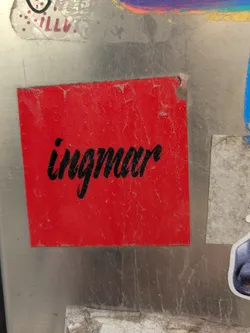 Street sticker The central sticker is a vibrant red square with the name 'ingmar' printed in black cursive script. The sticker appears old, with some wear and tear evident around the edges. The background features other stickers and a surface that shows signs of age and use.