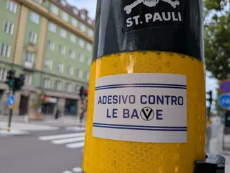 Street sticker A rectangular white sticker with a double blue horizontal border at the top and bottom. The text 'ADESIVO CONTRO LE BAVE' is written in a bold, blue, sans-serif typeface. The letter 'V' in 'BAVE' is enclosed in a circle with a small star at its top opening. The sticker is pasted onto a yellow reflective pole with a honeycomb pattern.