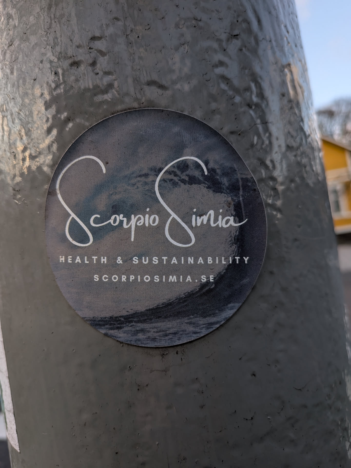 A circular sticker featuring a dark, textured background that evokes a stormy ocean or a deep, swirling wave, particularly prominent at the bottom. The brand name "Scorpio Simia" is elegantly displayed in a flowing white script font across the center. Below this, in a smaller, clean white sans-serif uppercase font, are the words "HEALTH & SUSTAINABILITY", followed by the website "SCORPIOSIMIA.SE" in the same style. The overall design is sophisticated and minimalist, with a strong focus on its brand identity and message.