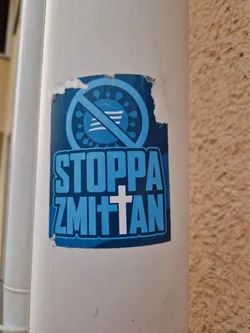 A blue sticker with a symbol of a virus with a cross on it and the text "Stoppa Zmitten". The sticker is slightly damaged.