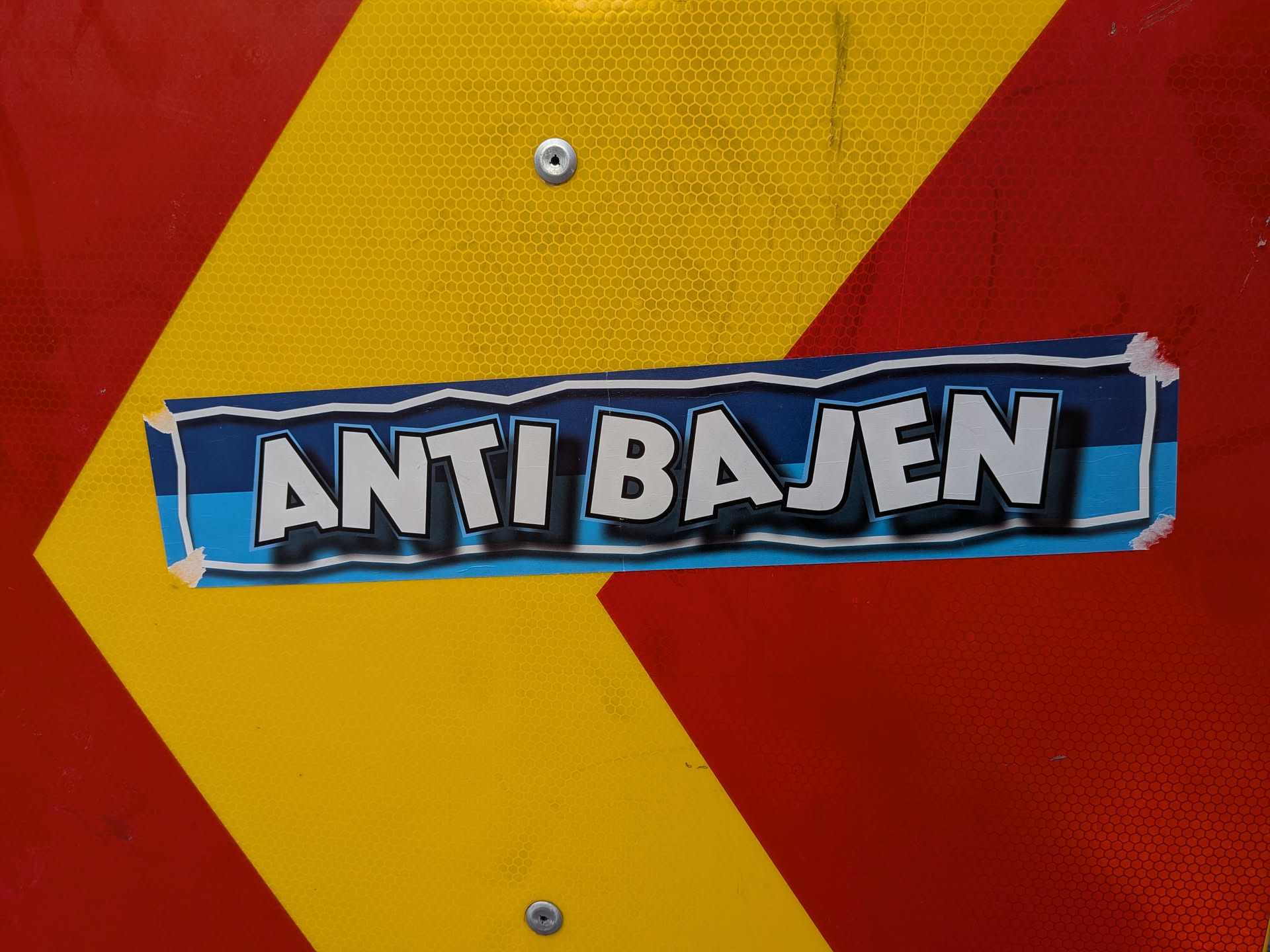 A rectangular sticker with 'ANTI BAJEN' in bold white letters with black shadows, set against a blue striped background. It is pasted onto a reflective red and yellow sign.