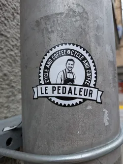 Street sticker A circular, black and white sticker featuring a smiling man with a beard, wearing a collared shirt, suspenders, and an apron. The man is centered within an inner white circle. Around the top half of this inner circle, the text 'CYCLE AND COFFEE' is repeated, separated by a small coffee bean icon. The outer edge of the sticker is a black border designed to resemble a bicycle gear or sprocket. A white banner with black text, 'LE PEDALEUR', stretches across the bottom of the central image, extending slightly beyond the main circle. The overall aesthetic is clean, illustrative, and has a vintage badge-like quality.