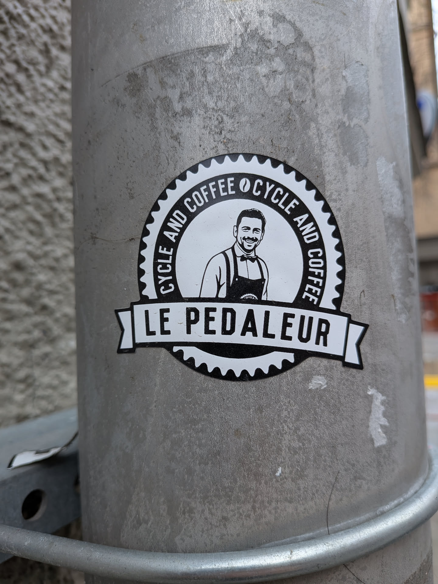 A circular, black and white sticker featuring a smiling man with a beard, wearing a collared shirt, suspenders, and an apron. The man is centered within an inner white circle. Around the top half of this inner circle, the text 'CYCLE AND COFFEE' is repeated, separated by a small coffee bean icon. The outer edge of the sticker is a black border designed to resemble a bicycle gear or sprocket. A white banner with black text, 'LE PEDALEUR', stretches across the bottom of the central image, extending slightly beyond the main circle. The overall aesthetic is clean, illustrative, and has a vintage badge-like quality.