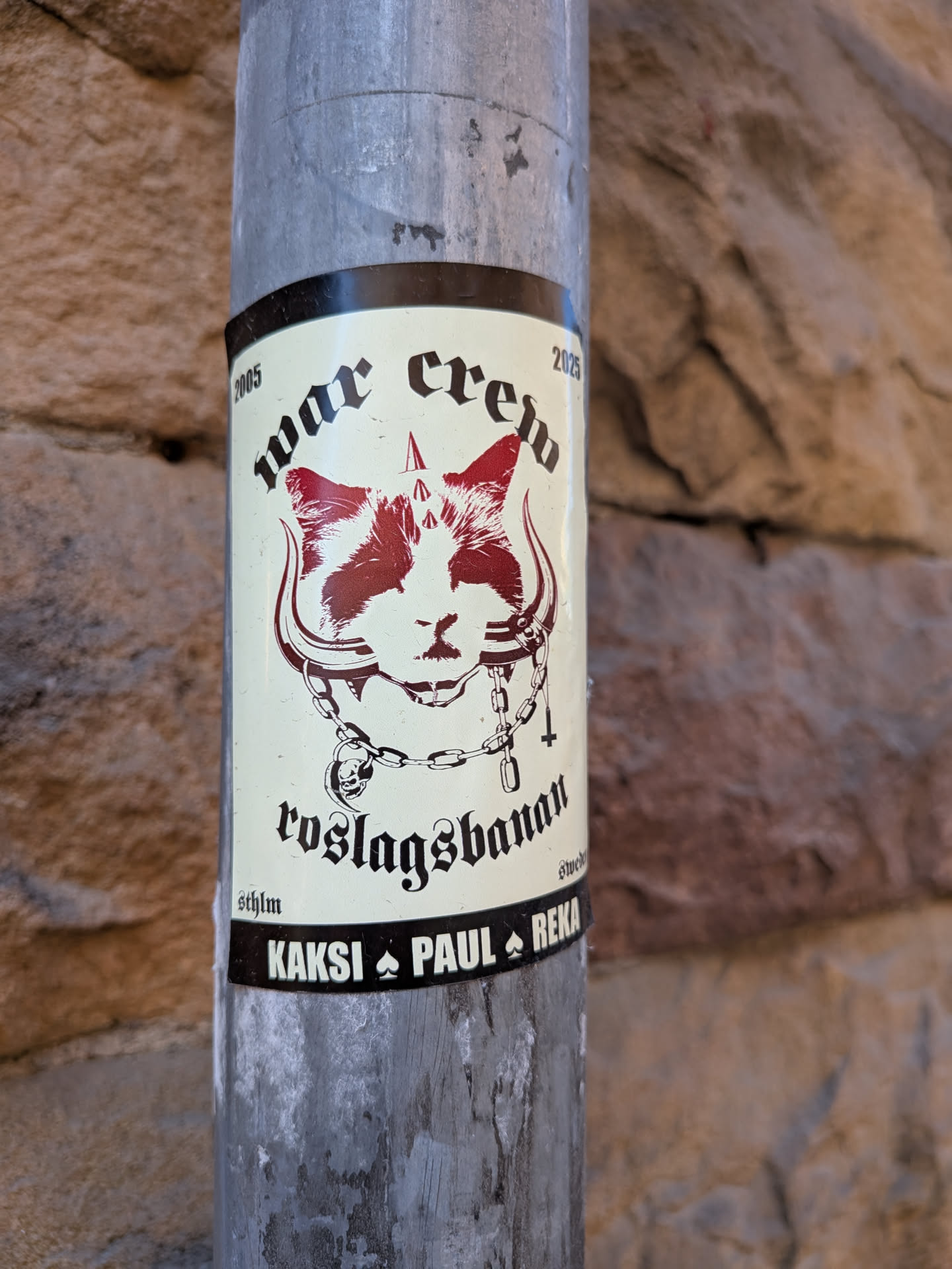 A rectangular sticker with a cream-colored background and a black border, affixed to a metal pole. The central image is a stylized cat's head rendered in dark red and white, featuring a stern expression. Above the cat's head are three small, dark red spikes. The cat wears a collar adorned with two large, curved horns extending outwards, resembling a bull's horns. A chain drapes below the cat's chin, from which a small skull charm hangs on the left and an inverted cross charm hangs on the right. The text 'war crew' is prominently displayed in a gothic font above the cat, flanked by '2005' on the left and '2025' on the right. Below the cat, 'roslagsbanan' is written in a similar gothic script. Smaller text 'sthlm' is on the bottom left and 'swede' (partially visible) on the bottom right. A black strip at the very bottom features the names 'KAKSI', 'PAUL', and 'REKA', separated by spade symbols.