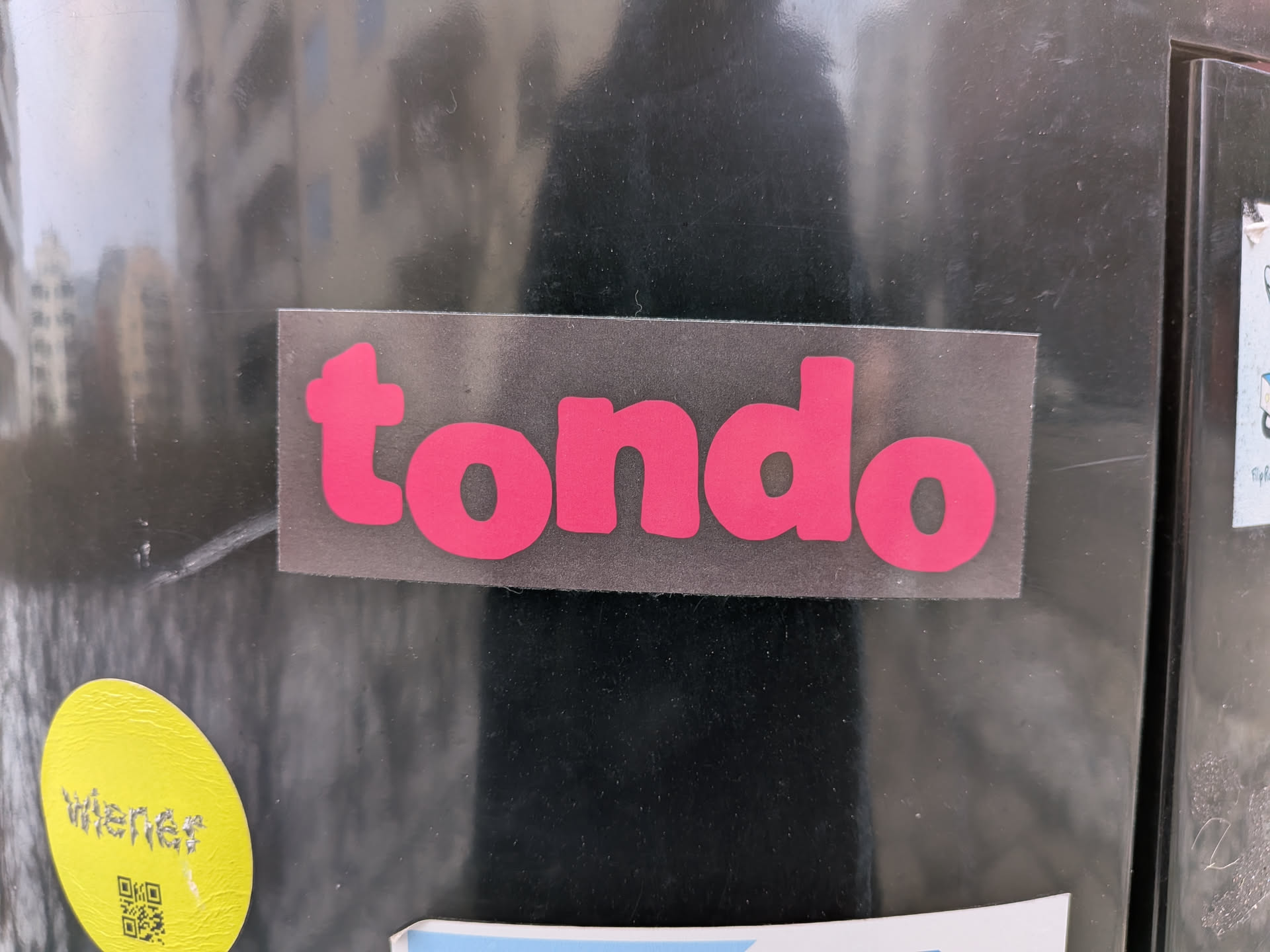 Street sticker The sticker is rectangular with slightly rounded corners, featuring the word 'tondo' in a bold, rounded, sans-serif font. The letters are a bright, saturated pink color, set against a dark grey or black background. The sticker appears to be applied to a reflective, dark surface, possibly a metal door or panel, with some minor wear and scratches visible on the sticker's surface and the surrounding area.