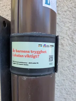 Street sticker The sticker is a rectangular design with a light green background, wrapped around a dark brown pipe. At the top left, there's a small RFSL logo featuring rainbow-colored bars. To its right, the RFSL Ungdom logo displays a rainbow circle, followed by 'RFSL UNGDOM STOCKHOLM' in black text. The central and most prominent text, in a bold red/pink font, asks 'Är barnens trygghet i skolan viktigt?' (Is children's safety in school important?). Below this, in smaller black text, it reads 'Sätt press på makthavarna – skriv under för trygghet och respekt i skolan.' (Put pressure on those in power – sign for safety and respect in school.). On the right side of the sticker, a black and white QR code is visible, accompanied by small black text underneath that says 'Scanna och skriv under!' (Scan and sign!). The sticker shows minor signs of wear and conforms to the curvature of the pipe.
