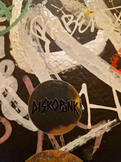 Street sticker A round, metallic gold sticker with the word "DISKOPUNK" in a bold, black, stylized font. It is affixed to a wall covered in various graffiti tags and paint splatters.