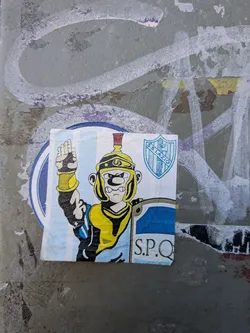 Street sticker The sticker features a cartoonish depiction of a Roman soldier in a yellow and black uniform with a red-plumed helmet. The soldier has a stern expression and is raising one hand in a salute. He is holding a shield with blue and white diagonal stripes, and the 'S.P.Q.' emblem is visible on the lower part of the shield. In the upper right corner of the sticker, there is a blue shield with white lettering that reads 'S.S.LAZIO'. The background of the sticker consists of light blue and white vertical stripes. The sticker appears to be slightly creased and adhered to a textured surface with some graffiti.